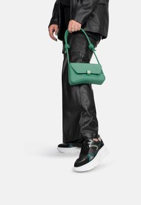 Borsa a tracolla verde in pelle con manico annodato, forma rettangolare, chiusura metallica. Abbinata a pantaloni in pelle neri e sneakers multicolori.