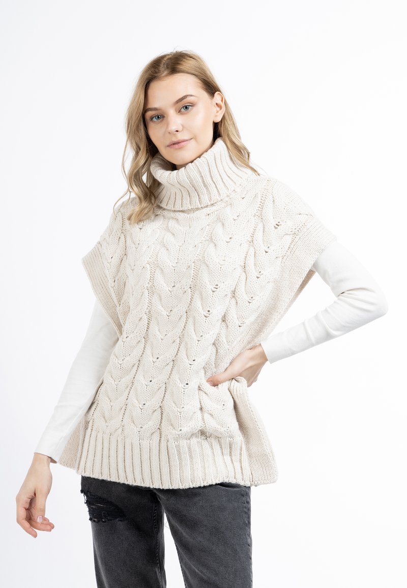 Beige gestrickter Poncho mit Zopfmuster und hohem Rippkragen. Verfügt über Seitentaschen und eine lockere Passform, kombiniert mit einem weißen Langarmshirt.