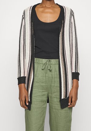 Osoba ubrana w otwarty, paskowany kardigan dziany z czarnymi, białymi i beżowymi paskami, na czarnym topie na ramiączkach i oliwkowozielonych spodniach z troczkami.