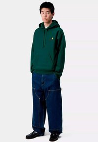 Sudadera verde con capucha y bolsillo frontal, combinada con jeans de pierna ancha azules. Zapatos negros completan el atuendo. Diseño simple y textura suave.