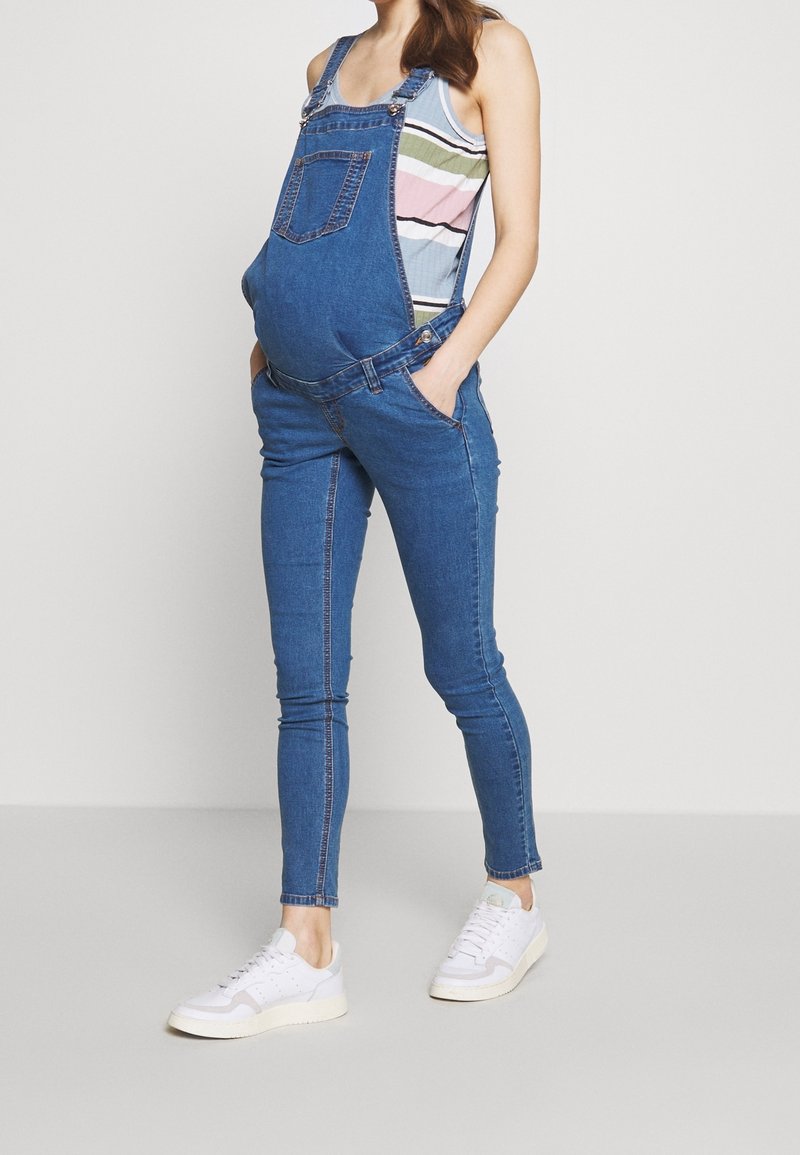 Femme enceinte portant une salopette de maternité en denim bleu, un débardeur rayé et des baskets blanches, debout les mains dans les poches.