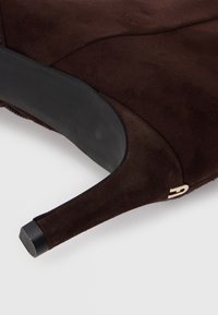 Brown suede high heel shoe with a sleek design, pointed toe, and gold hardware accent. The sole is black with a textured grip.
