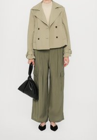 Veste croisée kaki, haut beige, pantalon large olive et chaussures plates noires ; tenant un sac à main noir noué.