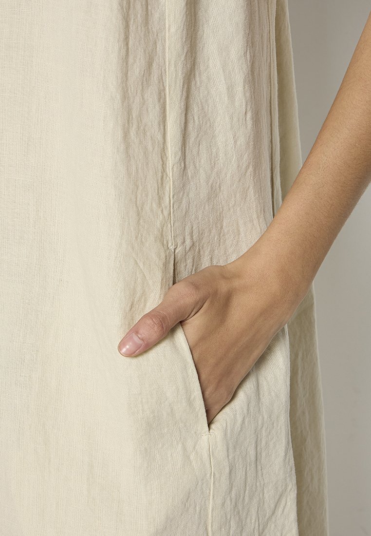 Helles beiges Kleidungsstück mit strukturiertem Finish, ausgestattet mit einer Seitentasche und einem lockeren Schnitt, teilweise gezeigt mit einer Hand, die in der Tasche ruht.