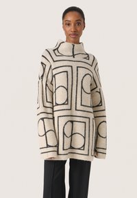 Beige tröja med svarta geometriska mönster, hög krage, oversized passform och texturerat stickat. Matchas med svarta raka byxor.