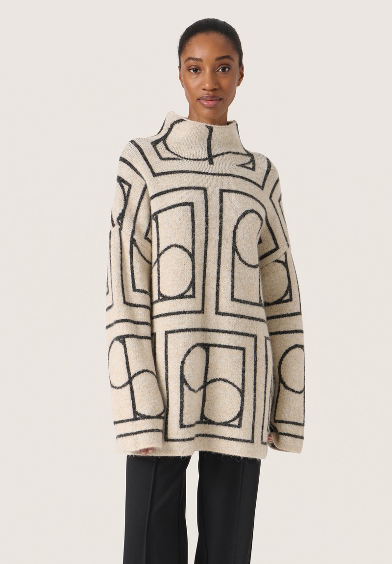 Beige tröja med svarta geometriska mönster, hög krage, oversized passform och texturerat stickat. Matchas med svarta raka byxor.