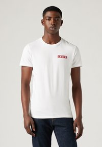 Tricou alb din bumbac, cu o etichetă roșie cu logo-ul Levi's pe pieptul stâng. Mâneci scurte, gât rotund și un design ajustat. Simplu și minimalist.