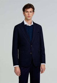 Blazer navy con dos bolsillos frontales, cierre de un solo botón y solapas con muesca. Llevado sobre un suéter azul y una camisa blanca con cuello.