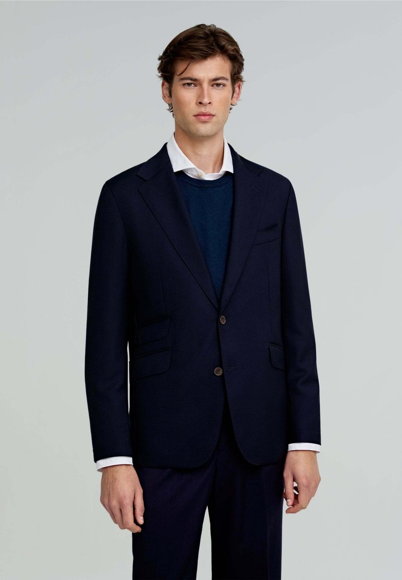 Blazer navy con dos bolsillos frontales, cierre de un solo botón y solapas con muesca. Llevado sobre un suéter azul y una camisa blanca con cuello.