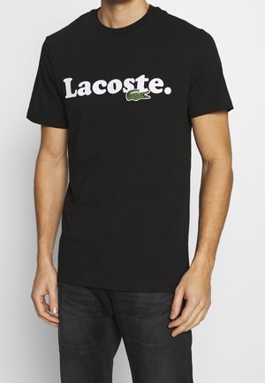 Homme portant un t-shirt noir avec le texte blanc "Lacoste." et le logo crocodile vert, associé à un jean foncé, sur fond blanc uni.