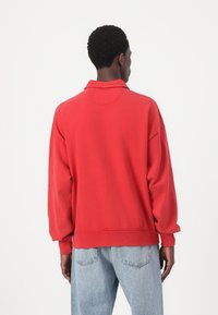 Felpa rossa con colletto classico, spalle scese e polsini a coste, abbinata a jeans in denim azzurro chiaro. Texture liscia, vestibilità rilassata.