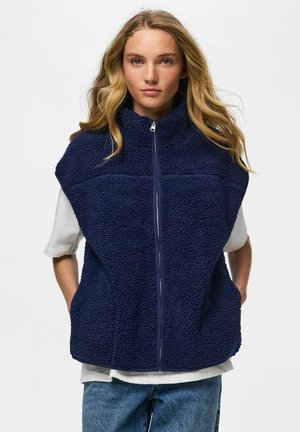 Femme blonde portant un gilet polaire bleu marine zippé par-dessus un t-shirt blanc et un jean bleu, debout les mains dans les poches devant un fond blanc.