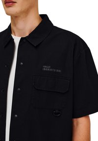 Zwart overhemd met korte mouwen en een knoopsluiting bij de kraag, met een voorzak, verdiept tekst en lusdetail. Gemaakt van stevig materiaal.