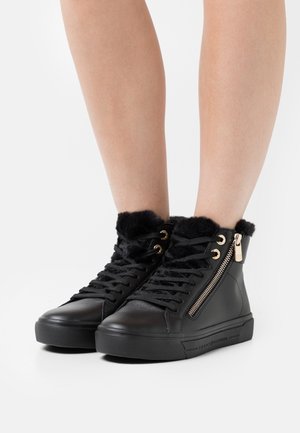 Mustat korkeavartiset lenkkarit turkisvuorella, sivuvetoketjulla ja nauhoilla, paljailla jaloilla valkoista taustaa vasten.