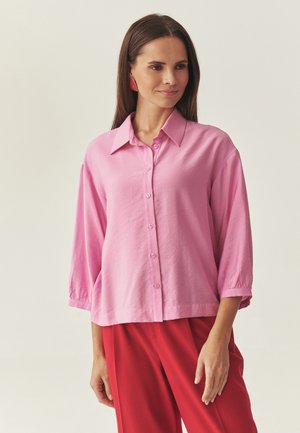 Różowa koszula na guziki z kołnierzykiem, rękawami o długości do łokcia i luźnym krojem, wykonana z lekkiego materiału. W połączeniu z czerwonymi spodniami.