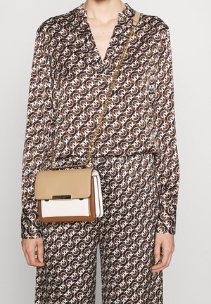 Vrouw draagt een bruin bloes met lang mouwen en een patroon, en bijpassende broek, ze draagt een kleine beige, witte en bruine handtas met een gouden kettingriem.