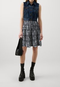 Džínová vesta bez rukávů s přiléhavým střihem, kombinovaná s vícedílnými černo-bílými vzorovanými šaty, robustními černými platformovými botami a černou kabelkou.