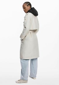 Long manteau beige avec une capuche matelassée noire, présentant une taille cintrée, des poches latérales et des poignets boutonnés. Porté par-dessus un jean bleu large.