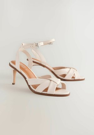 Beige hoge hakken sandalen met een slanke hak, met een gekruiste bandontwerp en een enkelband met een zilveren gesp. Gladde lederen textuur.