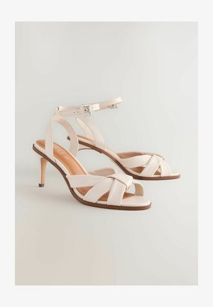 Beige High-Heelsandalen mit schmalem Absatz, die ein über Kreuz geführtes Riemendesign und einen Knöchelriemen mit einer silbernen Schnalle aufweisen. Glattes Leder.