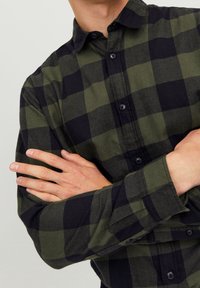 Camicia in flanella a quadri verde e nera con colletto button-down, maniche lunghe e bottoni scuri; il tessuto presenta una finitura soffice e testurizzata.