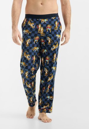 Hombre con pantalones de pijama azul marino estampados con guepardos sobre un fondo oscuro a cuadros, de pie descalzo frente a un fondo blanco.