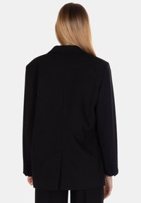 Blazer nero oversize con un design strutturato, revers a punta e uno spacco posteriore singolo. Il tessuto appare liscio con polsini abbottonati.