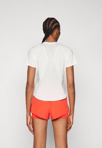 Camiseta deportiva blanca de manga corta, con un respaldo texturizado y dobladillo redondeado, combinada con pantalones cortos naranjas que presentan un diseño en capas y acentos reflectantes.