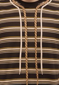 Κοντινό πλάνο καφέ ριγέ πουκάμισου με οριζόντιες λευκές, μπεζ και κίτρινες γραμμές και δύο ζευγάρια κρεμασμένων κορδονιών στη λαιμόκοψη.