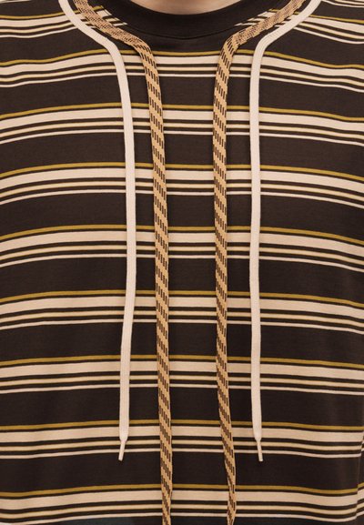 Gros plan sur une chemise marron à rayures avec des lignes horizontales blanches, beiges et jaunes, et deux paires de cordons pendants au niveau du col.