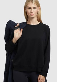 Black sweatshirt with a crew neck and raglan sleeves, made of soft fabric. The individual holds a navy puffer jacket over one shoulder.