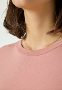 Koszula z krótkim rękawem w kolorze różowym, z okrągłym dekoltem, wykonana z miękkiej tkaniny. Widoczny jest delikatny naszyjnik z cienkiego złotego łańcuszka.