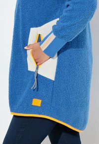 Blaues Fleece-Pullover mit einer weißen Brusttasche und orangefarbenen Akzenten, ausgestattet mit einem Reißverschluss und einem strukturierten Stoff.