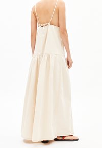 Beigeistä puuvillasta valmistettu maksi-mekko, jossa on vartalonmyötäinen yläosa, ohuet olkaimet ja kerroksittain laskeutuva, virtaviivainen helma. Mekossa on myös takaosan solmittava yksityiskohta.