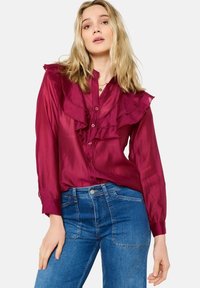 Burgundy blouse with ruffled front, long sleeves, and button closure. Worn with blue denim jeans, featuring a fitted design and pockets.
