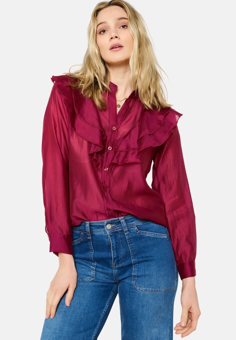 Burgundy blouse with ruffled front, long sleeves, and button closure. Worn with blue denim jeans, featuring a fitted design and pockets.