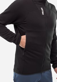 Veste en polaire noire avec une fermeture éclair à l'avant, présentant un matériau texturé, des coutures angulaires et des poches latérales pour un usage pratique.