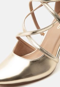 Γυναικεία χρυσή μεταλλιζέ παπούτσια με μυτερή μύτη, λουράκια που διασταυρώνονται στον αστράγαλο και λεία επιφάνεια.