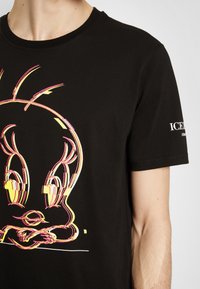 T-shirt noir avec un graphisme coloré et stylisé du visage de Titi et un texte blanc sur la manche droite.