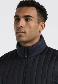 Marineblaue gefütterte Jacke mit hohem Kragen, vertikalem Steppmuster und Reißverschluss. Verfügt über ein kleines Pinguin-Logo auf der linken Seite.