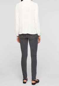 Blusa bianca a costine con maniche lunghe, caratterizzata da un orlo svasato. Abbinata a jeans attillati grigio scuro e scarpe nere flat.