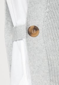 Un gilet tricoté gris clair présente un motif texturé, avec une ceinture en tissu et un bouton rond marron accentuant le détail sur le côté.