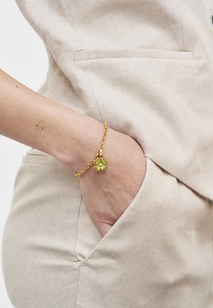 Roka v žepu bež obleke, ki nosi zapestnico z zlato verižico in obeskom iz zelene štirisolistne deteljice.