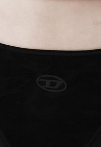 Reggiseno sportivo nero con un pattern testurizzato, dotato di un'etichetta rotonda nera con logo. Il design include una morbida fascia elastica lungo i bordi.