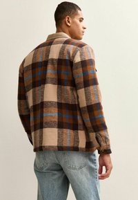 Brown plaid jacket made of textured fabric featuring blue and cream squares, with a button detail on the cuff and a wearable collar.