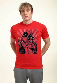 Červené bavlněné tričko s grafickým potiskem postavy, která vytváří tvar srdce. Design zahrnuje silné černé kontury a barevné kontrasty ve stylu komiksu.