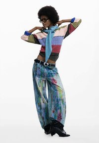 Pull tricoté coloré avec des textures variées, associé à un jean à jambes larges orné d'un motif éclaboussé de peinture. Des bottines noires à talons complètent la tenue.