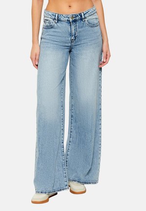 Mujer que lleva vaqueros de pierna ancha de talle alto en azul claro con botón y cremallera delanteros, combinados con zapatos blancos con suela de goma.