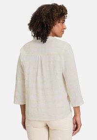 Hellbeige Bluse mit Dreiviertelärmeln, die aus einem strukturierten Stoff gefertigt ist und farbenfrohe Stickereien in Linien aus rosa, grün und orangefarbenen Punkten aufweist.