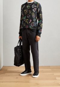 Person, die einen schwarzen weihnachtlich gestalteten Pullover mit bunten Feiertagsmotiven trägt, schwarze Hosen, schwarze Sneaker mit weißen Sohlen, und eine schwarze Tasche hält.